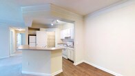 This single-story space features an open-concept layout that seamlessly connects the living area and kitchen through a wide arched passageway. The kitchen is equipped with white cabinetry, a white electric range, and an under-cabinet range hood, all set against light-toned countertops and a subtle tile backsplash. Recessed track lighting illuminates the kitchen, enhancing its clean, functional aesthetic. A peninsula-style breakfast bar extends from the kitchen, providing a natural transition into the adjacent living area and offering additional counter space for dining or casual use. The living area is finished with warm hardwood-style flooring that carries throughout, adding visual continuity across the space. Crown molding lines the ceilings, lending a refined architectural detail throughout. The walls are painted in a soft, neutral cream tone, creating a bright and welcoming ambiance. A door visible near the kitchen leads to what appears to be a pantry or storage closet, adding practical utility. Overall, the space presents a clean, well-maintained interior with a cohesive and functional design.