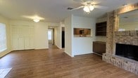 This single-story home offers a well-connected layout with a warm, functional design throughout. The main living area is anchored by rich hardwood-style flooring that extends seamlessly through the space, complemented by a ceiling fan with integrated lighting that ensures comfort and brightness. A defining architectural feature is the pass-through window connecting the living area to the kitchen, fostering an open, communicative flow between the two spaces. The kitchen is equipped with dark wood cabinetry, a built-in oven, and a refrigerator, providing ample storage and utility. Adjacent to the kitchen pass-through, a built-in shelving unit with dark wood cabinetry sits alongside a stone or brick accent detail, adding a touch of character and texture to the room. A hallway visible from the main living area leads toward additional rooms, hinting at a practical and thoughtfully arranged floor plan. The home's interior palette of white walls and neutral tones creates a clean, inviting atmosphere, while the overall layout maximizes usability and ease of movement throughout the single floor.