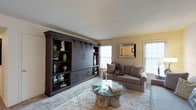 Westbury Harbor is a thoughtfully designed single-floor residence that offers a well-rounded combination of comfort and functionality across its living spaces. The living room serves as a welcoming centerpiece, featuring a striking dark wood built-in wall unit with barn-style sliding doors and open shelving, complemented by plush neutral sofas, a distinctive glass-top coffee table with a natural wood base, and multiple windows that invite natural light into the warm, carpeted space. The dining area accommodates generous group gatherings with a large white table surrounded by dark spindle-back chairs, accented by a classic wall clock and adjacent display shelving. The kitchen is outfitted with white cabinetry offering abundant storage, a full suite of white appliances including a range, refrigerator, and dishwasher, and a parquet-style floor that adds character. The bedrooms are carpeted and offer natural light through draped windows, with the primary bedroom featuring dual reach-in closets and the second bedroom providing additional closet storage. Two bathrooms are finished with white tile wainscoting and warm-toned vanity countertops; one features coastal-themed décor and the other a patterned shower curtain and tiled flooring. Throughout, the residence maintains a cohesive, neutral palette with clean lines and practical design.