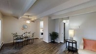 This single-floor space showcases an open-concept layout that connects the living, dining, and kitchen areas in a cohesive and functional design. Dark hardwood-style flooring runs throughout, creating a bold contrast against the light gray walls and white trim. Exposed structural beams add architectural character to the textured ceiling, while a ceiling fan with a light fixture provides both practicality and a subtle design element.

The dining area is defined by a round glass-top table paired with elegant wrought-iron chairs, anchored by a decorative sunburst mirror on the wall. A potted fern placed near the central partition brings a natural touch to the space. The kitchen, visible through an open pass-through, features white cabinetry and white appliances, maintaining a clean, bright aesthetic.

Two white doors are visible toward the rear of the space, suggesting access to additional rooms or storage, one of which appears to lead to a walk-in closet or utility area. A console table with a contemporary lamp rounds out the living area. Overall, the space presents a clean, well-organized interior with a neutral palette and practical design.
