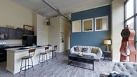 This single-floor space showcases a thoughtfully designed open-concept layout that seamlessly connects the kitchen and living areas. The kitchen features a sleek breakfast bar with industrial-style bar stools, dark countertops, and modern appliances, making it an ideal spot for casual dining or entertaining. Exposed ductwork runs along the high ceilings, lending the space a distinct industrial character that complements the contemporary aesthetic throughout.

The living area is anchored by a striking deep blue accent wall adorned with three white-framed pass-through windows, adding architectural interest and visual depth. Light hardwood-style flooring extends throughout, creating a cohesive and airy feel. A tufted gray sofa paired with a dark coffee table serves as the centerpiece of the lounge area, while a built-in shelving nook along one wall offers functional display space. Neutral walls balance the bold accent tones, and the overall palette of whites, grays, and blues gives the space a modern yet approachable ambiance. The generous ceiling height and open floor plan contribute to a notably spacious and well-proportioned interior.