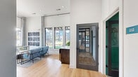 Located at 112 Point Pl in Mooresville, NC, this thoughtfully designed single-floor space serves as a vibrant community lounge and workspace. The interior blends coastal-inspired aesthetics with modern sophistication through a cohesive palette of teal, warm gold, and crisp white tones throughout.

The living area is the heart of the space, boasting soaring ceilings that amplify its openness. Large floor-to-ceiling windows flood the room with natural light and offer views of an outdoor pool area. Rich light oak hardwood flooring runs throughout, complementing plush blue velvet seating arranged around a marble-topped coffee table. A striking textured wave-panel accent wall in teal frames a dining nook outfitted with tan leather booth seating, dark bistro tables, and elegant gold pendant lighting.

The two office spaces offer a more focused atmosphere. Each features a sleek marble-surface desk, a cognac leather chair, and refined geometric patterned wallcovering, creating a calm and productive environment. Soft overhead pendant lighting and lush greenery add warmth to each workstation.

Curated artwork, including a bold grid of abstract panels, adds a gallery-like character to the lounge. The seamless blend of functional zones and design-forward details makes this space both practical and visually compelling.