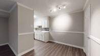 This single-story residence offers a well-organized layout with distinct living, dining, kitchen, and sleeping areas connected by wood-look laminate flooring in neutral tones. The open living and dining space features crown molding, a textured ceiling, and track lighting that illuminates the area effectively. A partial wall divides the dining space from the rest of the unit, adding architectural definition while preserving an open feel.

The galley-style kitchen is efficiently designed with white shaker cabinetry, marble-look countertops, and a mosaic tile backsplash in a neutral black-and-white pattern. Black appliances, including a range and refrigerator, complement the clean aesthetic, while a stainless steel sink with a gooseneck faucet and dishwasher add functional convenience. Under-cabinet lighting and an overhead exhaust hood complete the kitchen.

The two bedrooms are carpeted and each include a ceiling fan with light fixture. One bedroom benefits from a window that allows natural light. Two bathrooms serve the unit — one features a shower-tub combination with a curtain rod, white vanity, and wood-look flooring, while the second offers a vanity with lighted mirror and similar flooring. The overall palette is neutral throughout, creating a cohesive and understated ambiance.
