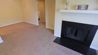 This single-floor space presents a warm and understated aesthetic characterized by neutral tones throughout. The living area is anchored by a classic fireplace framed with a clean white mantel and a dark hearth surround, serving as a natural focal point within the room. Plush wall-to-wall carpeting in a soft, neutral hue extends across the floor, contributing to the overall sense of comfort and warmth. The walls are finished in a light beige tone, creating a bright and inviting atmosphere. White baseboards provide a crisp contrast along the perimeter of the room. A hallway visible from the living area leads to additional spaces, including what appears to be a bathroom, suggesting a thoughtfully connected layout. The space maintains a simple, functional design with clean lines and minimal ornamentation, making it highly adaptable to a variety of interior styles. The overall ambiance is calm and livable, well-suited for everyday comfort.