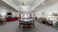 Located on Blue Heron Boulevard in Fond du Lac, Wisconsin, this single-story community facility offers a thoughtfully designed layout with a variety of functional and inviting spaces. The main gathering area is a standout feature, showcasing tray ceilings adorned with elegant brass chandeliers, recessed lighting, and plush carpeting throughout. Multiple round tables with cushioned roller chairs provide ample seating for group activities and socializing, while framed nature and wildlife artwork lends a warm, classic aesthetic to the walls.

A dedicated lounge area offers a more relaxed setting, centered around a stone-surround fireplace topped with a wall-mounted television, complemented by generous red upholstered seating and decorative accents. Adjacent seating nooks with striped armchairs and wood-trimmed interior windows provide quieter spaces for individual relaxation.

The entrance foyer is bright and welcoming, with large exterior windows offering views of the parking area and natural light throughout. Wood-trimmed doorways and warm neutral tones create a cohesive, comfortable atmosphere across all areas.

A well-equipped fitness room rounds out the amenities, featuring cardio machines, a multi-station cable system, free weights, adjustable benches, wall mirrors, and large windows that fill the space with natural light — making it a practical and pleasant environment for exercise.