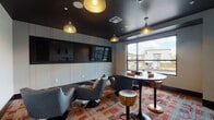 Located on SW 175th Ave in Beaverton, Oregon, this single-floor communal lounge space offers a thoughtfully designed environment built for social gathering and relaxed interaction. The room is defined by a striking contrast between its dark, matte ceiling and crisp white walls, creating a modern and intimate atmosphere. Bold pendant lights with gold interiors hang from above, casting a warm, inviting glow throughout the space.

The lounge is furnished with a mix of contemporary seating arrangements, including plush swivel armchairs in cool grey upholstery accented with decorative pillows, paired alongside sleek round tables in both low and high-top configurations. Wooden accent stools add a natural warmth that balances the room's modern aesthetic. A vibrant, multi-toned patterned carpet in red, grey, and neutral tones anchors the space and adds visual energy underfoot.

A wall-mounted flat-screen TV is integrated into a paneled feature wall, making the room well-suited for viewing or entertainment. A large window allows natural light to filter in and provides views of the surrounding Beaverton neighborhood. Multiple entry doors, audio controls, and thoughtful built-in cabinetry complete the functional design. Overall, the space presents a polished, amenity-focused lounge ideal for community use.