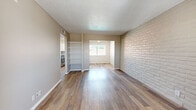 This single-floor residence offers a well-organized layout with a blend of practical design elements and distinctive architectural character. The living area serves as the heart of the home, showcasing warm wood-look flooring that extends throughout the main spaces, a textured popcorn ceiling, and a striking white-painted brick accent wall that adds texture and visual interest. Built-in open shelving provides convenient storage and display space, while a pass-through opening connects the living area to an adjacent dining or secondary living space, both bathed in natural light from windows overlooking the outdoors. The kitchen is accessible from the main living area and features a stainless steel range visible from the adjoining space. The bedrooms are carpeted for comfort, each featuring ceiling fans with light fixtures, white-painted brick walls, and windows that allow natural light to filter in. The bathroom is well-appointed with wood-look flooring, a white vanity with a speckled countertop, a large mirror, a shower-tub combination, and a white-painted brick wall that ties the home's design language together cohesively. Neutral, warm-toned walls throughout the residence create a clean, move-in-ready atmosphere.