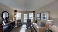 This single-floor space centers around a thoughtfully designed living room that combines comfort and style. The room is finished in warm neutral tones, with beige walls and light-colored carpeting that create a cohesive, inviting atmosphere. Natural light pours in through multiple large windows, each dressed with decorative curtains featuring a bold, wavy pattern in contrasting dark and light tones, adding a touch of visual interest to the space.

A generously sized cream-colored sofa anchors the seating area, accented with patterned throw pillows in shades of blue and grey. Two grey upholstered armchairs are positioned across from the sofa, creating a balanced and functional conversation area. A rectangular coffee table serves as the centerpiece of the arrangement, adding both practicality and style.

A glass-paneled door provides access to an outdoor area, further enhancing the room's connection to natural light and the exterior surroundings. Framed artwork displayed on the walls contributes to the polished, finished look of the space. A flat-screen television on a dark media console completes the room's functional offerings, making this living area both stylish and livable.