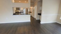 This single-floor space showcases an open-concept design that integrates the kitchen and living areas into a cohesive, functional layout. The kitchen is appointed with white cabinetry, dark granite countertops, and stainless steel appliances, including a gas range, microwave, and refrigerator. A peninsula with matching dark countertop extends into the open living space, offering additional prep and seating potential while defining the kitchen zone without enclosing it. Dark hardwood flooring runs throughout, providing a rich contrast against the neutral, light-toned walls and white trim. Recessed lighting in the kitchen area ensures practical illumination, while the open ceiling height adds a sense of spaciousness. A hallway visible beyond the kitchen leads to additional rooms, suggesting a well-organized floor plan. A secondary counter nook is visible to the right, potentially serving as a bar or additional workspace. The overall aesthetic is clean and contemporary, with a neutral palette that allows for versatile interior styling.