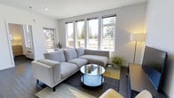 This single-floor residence showcases a clean, contemporary design with a strong emphasis on natural light and modern simplicity. The living area serves as the heart of the home, anchored by a generous bank of large casement windows that frame views of mature trees and the surrounding landscape, flooding the space with abundant daylight. The neutral white walls and dark flooring create a crisp contrast that enhances the bright, airy atmosphere throughout.

The living room is thoughtfully appointed with a low-profile sectional sofa in a soft gray upholstery, paired with a round glass-top coffee table that adds a light, open feel to the seating area. A layered area rug in muted tones of gray, yellow, and blue grounds the space with a subtle pop of color. Potted greenery introduces a natural, refreshing element to the interior. An additional side window allows natural light to enter from multiple angles, further brightening the room.

The overall ambiance is calm and inviting, with a minimalist sensibility that keeps the space feeling uncluttered and functional. The modern architectural detailing, recessed lighting, and thoughtful layout reflect a well-considered design approach suited to contemporary living.