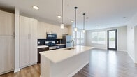 Located at 3200 Dr Andrew J Brown Ave in Indianapolis, IN, this modern single-floor unit offers a thoughtfully designed open-concept layout. The combined kitchen and living area serves as the heart of the space, showcasing a large quartz-topped kitchen island with an integrated sink, illuminated by a series of sleek pendant lights. The kitchen is outfitted with stainless steel appliances — including a refrigerator, range, and microwave — complemented by two-tone cabinetry featuring light wood upper cabinets and dark lower cabinets, set against a dark tile backsplash. Tall pantry-style cabinetry provides generous storage. The living area flows naturally from the kitchen, bathed in natural light from large windows with horizontal blinds and access to an exterior door. Light hardwood-style flooring runs throughout the entire unit, adding warmth and visual continuity. The bedroom is a calm, neutral-toned retreat with a large window offering outdoor views, direct access to the bathroom, and a dedicated closet. The bathroom features a full soaking tub with shower, a dark navy vanity with white countertop, and hardwood-style flooring, creating a clean, contemporary finish. The overall ambiance is bright, modern, and efficiently organized.