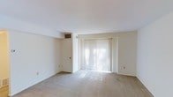 This single-floor space presents a straightforward and functional layout centered around a spacious, open living area. The room is finished with neutral-toned wall-to-wall carpeting that extends throughout, providing a soft underfoot feel and a cohesive, understated aesthetic. White walls contribute to a clean, bright atmosphere throughout the space.

A prominent feature of the living area is the large sliding glass door dressed with vertical blinds, which allows generous natural light to filter into the room, casting soft, diffused illumination across the floor. The door provides potential access to an outdoor area and serves as a key source of daylight. A standard door with an overhead HVAC vent is positioned along the adjacent wall, indicating a dedicated climate control system for year-round comfort.

The ceiling is smooth and flat, maintaining the room's simple, uncluttered character. The overall layout is open and versatile, offering ample flexibility for various furniture arrangements. The neutral finishes and minimal detailing create a blank canvas well-suited for a range of interior styles, making this a practical and adaptable living space.