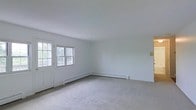 This single-floor space presents a straightforward and functional layout designed for comfortable everyday living. The main living area is immediately welcoming, characterized by its light, neutral color palette with white walls and a smooth white ceiling that enhances the sense of openness and brightness. Soft, wall-to-wall carpeting in a muted gray tone covers the floor, adding warmth and a quiet, understated aesthetic to the room. Natural light enters through a pair of windows positioned alongside a paneled entry door, framing views of surrounding greenery and contributing to the airy atmosphere of the space. Baseboard heating units run along the lower perimeter of the walls, providing an efficient and unobtrusive heating solution well-suited to colder climates. The clean lines and minimal detailing of the interior create a versatile, move-in-ready environment that can be easily personalized. The overall ambiance is calm and functional, offering a blank canvas that is both practical and inviting for a variety of uses.