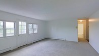 This single-floor space presents a straightforward and functional layout designed for comfortable everyday living. The main living area is immediately welcoming, characterized by its light, neutral color palette with white walls and a smooth white ceiling that enhances the sense of openness and brightness. Soft, wall-to-wall carpeting in a muted gray tone covers the floor, adding warmth and a quiet, understated aesthetic to the room. Natural light enters through a pair of windows positioned alongside a paneled entry door, framing views of surrounding greenery and contributing to the airy atmosphere of the space. Baseboard heating units run along the lower perimeter of the walls, providing an efficient and unobtrusive heating solution well-suited to colder climates. The clean lines and minimal detailing of the interior create a versatile, move-in-ready environment that can be easily personalized. The overall ambiance is calm and functional, offering a blank canvas that is both practical and inviting for a variety of uses.