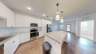 Located at 2280 Griffon Rd in Vero Beach, Florida, this efficiently designed one-bedroom, one-bathroom residence offers a clean, modern aesthetic throughout its single-floor layout. The open-concept kitchen and living area serves as the heart of the home, featuring warm wood-look flooring, crisp white walls, and abundant natural light from windows and a glass exterior door that offer views of the surrounding greenery. The kitchen is well-equipped with white shaker cabinetry accented by gold hardware, light granite countertops, a central island with pendant lighting, and a full suite of stainless steel appliances including a refrigerator, range, microwave, and dishwasher. The living area flows seamlessly from the kitchen, providing a versatile and bright open space. The bedroom is carpeted for comfort and features two windows that bring in ample natural light and outdoor views. It connects directly to a full bathroom finished with a granite-top vanity, a mirror, and wood-look flooring. A dedicated walk-in closet with built-in wire shelving provides practical storage. The residence presents a fresh, contemporary feel with consistent finishes and a functional, well-organized floor plan.