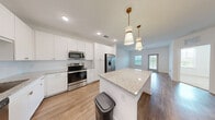 Located at 2280 Griffon Rd in Vero Beach, Florida, this efficiently designed one-bedroom, one-bathroom residence offers a clean, modern aesthetic throughout its single-floor layout. The open-concept kitchen and living area serves as the heart of the home, featuring warm wood-look flooring, crisp white walls, and abundant natural light from windows and a glass exterior door that offer views of the surrounding greenery. The kitchen is well-equipped with white shaker cabinetry accented by gold hardware, light granite countertops, a central island with pendant lighting, and a full suite of stainless steel appliances including a refrigerator, range, microwave, and dishwasher. The living area flows seamlessly from the kitchen, providing a versatile and bright open space. The bedroom is carpeted for comfort and features two windows that bring in ample natural light and outdoor views. It connects directly to a full bathroom finished with a granite-top vanity, a mirror, and wood-look flooring. A dedicated walk-in closet with built-in wire shelving provides practical storage. The residence presents a fresh, contemporary feel with consistent finishes and a functional, well-organized floor plan.