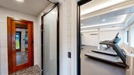 This gym and sauna facility offers a thoughtfully designed, single-floor fitness space that combines functionality with a clean, polished aesthetic. The exercise room serves as the centerpiece, featuring durable rubber flooring, white shiplap-accented walls, and a tray ceiling with recessed and ambient lighting that creates a bright, energizing atmosphere. Large windows allow natural light to filter in, enhancing the open feel of the workout area. The room is well-appointed with a range of cardio equipment including treadmills and stationary bikes, as well as strength-training equipment such as a multi-station cable machine, free weights, and benches. A wall-mounted TV, mirrored wall section, and a wooden storage shelf add both utility and visual appeal. Structural columns with classic detailing contribute an architectural touch to the space. A dedicated entrance with glass-panel doors provides seamless access, and a water fountain station is conveniently located nearby. The bathroom features a generously sized walk-in shower finished in large-format white marble-look tiles and grey mosaic accents, with a teak bench and frameless glass partition — delivering a spa-like, polished finish that complements the overall wellness focus of the facility.