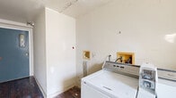 This single-room utility space serves as a dedicated laundry and utility area, currently undergoing renovation. The room features dark hardwood flooring throughout, which contrasts with the white-painted walls. A washer and dryer are present, with visible plumbing hookups recessed into the wall, indicating established laundry infrastructure. A notable feature is the bank of multiple utility meters mounted along one wall, suggesting this space serves a multi-unit building. An electrical panel is also mounted on the wall near a window, which allows natural light to enter and offers a view of the surrounding neighborhood. The entry is marked by a solid blue door with a small window, providing access to the corridor. The walls show signs of ongoing renovation work, including areas of exposed framing, drywall patches, and sections awaiting finishing. The ceiling also shows wear consistent with a space mid-renovation. Overall, this utilitarian space is structurally intact and presents a clear opportunity for a functional, finished laundry and utility room upon completion of the renovation work.
