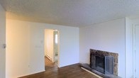 This single-floor space presents a straightforward and functional layout centered around a living area with notable character. The room features rich, dark hardwood flooring that extends throughout, adding warmth and a sense of continuity to the space. A stone-surround fireplace anchors one wall, providing a focal point and a cozy atmosphere. The textured popcorn ceiling and crisp white walls keep the space feeling clean and neutral, offering a versatile backdrop for various interior styles. A doorway leads to an adjacent room, visible in the background, which benefits from natural light filtering through a window — suggesting the layout flows comfortably from one area to the next. A baseboard heating unit is visible in the adjoining room, indicating a practical heating setup throughout. The overall ambiance is understated and move-in ready, with the combination of hardwood floors, a classic fireplace, and neutral finishes creating a livable, adaptable environment suitable for a range of furnishing styles and personal tastes.
