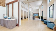 This single-floor commercial space presents a sophisticated, contemporary design centered around an expansive open lobby. Floor-to-ceiling glass walls and oversized gridded windows flood the interior with natural light, offering views of mature trees and outdoor plazas beyond. Light-toned large-format tile flooring extends throughout, enhancing the sense of openness and continuity across zones.

The main entry features glass double doors framed in dark metal, welcoming visitors into a bright reception area furnished with teal upholstered benches and dark accent seating. Soaring ceiling heights, structural white columns, and recessed track lighting define the architectural character of the space.

Moving deeper into the interior, a dedicated refreshment station displays a coffee machine set against a striking blue stone tile backsplash, complemented by warm wood cabinetry and white quartz countertops. Adjacent is a glass-enclosed conference room furnished with executive seating, visible from the main corridor.

Lounge areas are thoughtfully positioned near large windows, furnished with blue upholstered chairs, round ottomans, and sleek side tables. Decorative touches including sculptural glass art pieces and colorful vases add personality throughout. The overall ambiance is polished, functional, and welcoming.