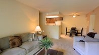 This single-floor residence offers an open and functional layout that connects the living, dining, and kitchen areas in one cohesive space. The living area is carpeted and comfortably sized, providing a relaxed setting anchored by neutral-toned walls that lend a warm, inviting ambiance throughout. A ceiling fan overhead serves both the living and dining zones, contributing to the room's practicality.

The kitchen is positioned at the rear of the main living space and is accessible via a raised pass-through counter, which creates a natural visual and functional connection between the kitchen and the surrounding areas without fully enclosing the space. Warm wood-toned upper cabinetry lines the kitchen walls, and a white refrigerator is visible, contributing to a classic, clean aesthetic. The pass-through counter also allows for casual interaction between the kitchen and adjoining spaces.

Adjacent to the kitchen, a dining area accommodates a small table and chairs, making it well-suited for everyday meals. The consistent warm wall tones and soft carpet throughout the main living spaces create a unified, cohesive interior that feels comfortable and move-in ready.