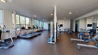 Located at 21834 Pacific Hwy S in Des Moines, Washington, this fitness center offers a generously sized, single-floor layout designed to accommodate a comprehensive range of workout needs. The space features durable rubber flooring throughout, providing both safety and comfort during exercise.

The cardio zone is prominently positioned along a wall of large, floor-to-ceiling windows that flood the space with natural light and offer pleasant views of the surrounding tree-lined landscape. Multiple treadmills, elliptical trainers, and a row of upright and recumbent cycling bikes—some equipped with interactive screens—make up this section of the facility.

A dedicated strength training area includes a cable multi-functional trainer, free weights, dumbbell racks, medicine balls, and a rowing machine. Full-length mirrors line one wall, enhancing the sense of space and allowing users to monitor their form during workouts. A wall-mounted television provides entertainment for those using nearby equipment.

The layout is thoughtfully organized with structural columns that naturally divide the space into distinct workout zones without disrupting the open, airy feel. Recessed ceiling lighting supplements the abundant natural light. The clean, modern aesthetic—characterized by white walls, high ceilings, and a neutral color palette—creates a professional and motivating environment well-suited for a broad range of fitness activities.