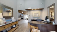 This well-appointed single-floor community space combines modern design with warm, inviting elements throughout its open-concept layout. Visitors are welcomed through a glass-paneled entrance foyer featuring a tufted leather accent chair, leading directly into a professional reception area with a dedicated workstation, recessed lighting beneath a floating soffit, and wood-look tile flooring that carries seamlessly throughout the space.

The expansive main lounge area commands attention with soaring ceilings, a dramatic gold-toned ceiling detail, and a striking floor-to-ceiling stacked stone fireplace topped with a wall-mounted flat-screen TV. Generous floor-to-ceiling windows along one wall flood the room with natural light and offer views of the surrounding landscaping. The lounge is furnished with leather seating, a cowhide area rug, and a wood coffee table, creating a relaxed yet sophisticated atmosphere.

Adjacent to the lounge, a dining area features a large wood table with upholstered chairs, illuminated by a mosaic tile backsplash and pendant lighting from the neighboring kitchenette. The kitchenette is equipped with rich wood cabinetry, granite countertops, a beverage refrigerator, and a central island with a sink. A quiet reading nook with a tufted armchair and dual windows rounds out this thoughtfully designed community space.