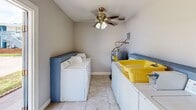 This single-floor laundromat is a compact, functional utility space designed specifically for laundry operations. The room is finished with neutral light gray walls and durable ceramic tile flooring, creating a clean and practical environment. Multiple top-load washing machines — including Whirlpool units — are arranged along the walls, accompanied by additional laundry appliances on the opposite side, maximizing the use of the available floor space. A standalone water heater with visible plumbing connections is situated in one corner, servicing the room's utility needs, alongside an electrical panel mounted on the wall nearby. A ceiling fan with a light fixture is centrally installed, providing ventilation and air circulation throughout the space. White baseboard trim lines the perimeter of the room, complementing the overall clean aesthetic. A door on one wall provides direct access to the exterior, allowing natural light to filter in and offering convenient entry and exit. A trash bin is also present, supporting day-to-day operational needs. Overall, the space is straightforward in its layout and purpose, efficiently accommodating essential laundry equipment within a well-maintained, utilitarian setting.