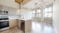 Located at 129 E Third St in downtown Dayton, Ohio, this one-bedroom, one-bathroom residence offers a thoughtfully designed layout across a single floor. The open-concept kitchen and living area serves as the heart of the unit, showcasing light wood cabinetry, white quartz countertops, a herringbone tile backsplash, and a central island with a brushed gold faucet. Stainless steel appliances, including a refrigerator, range, microwave, and dishwasher, complete the well-appointed kitchen. Pendant lighting with gold-toned fixtures adds a warm, modern touch, while exposed ductwork and a wood-paneled ceiling accent lend an industrial character to the space. Large windows in the living area flood the room with natural light and provide views of the surrounding cityscape. Light-toned wood plank flooring runs throughout, creating a cohesive and airy feel. A well-lit hallway with wall sconces leads to the private bedroom, which features neutral gray walls and the same continuous flooring. The bathroom is elegantly finished with large-format marble-look tile on both the floors and walls, a soaking tub, and a frameless sliding glass shower enclosure. Overall, the unit balances contemporary design with industrial charm in a prime downtown location.
