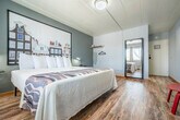 Located on East Washington Avenue in Madison, Wisconsin, this efficiently designed single-floor unit offers a self-contained layout with one bedroom and one bathroom. The main room serves as the primary living and sleeping space, finished with durable wood-look flooring and painted in a cool, neutral gray that lends the space a clean, modern feel. A queen-sized bed is the centerpiece of the room, complemented by a large framed wall art piece depicting European-style architecture, adding a decorative focal point. Natural light filters in through two windows dressed with curtain panels, offering views of the surrounding neighborhood. A wall-mounted flat-screen TV faces the bed, while a glass-top desk with seating provides a functional workspace. A compact mini-refrigerator and microwave are conveniently tucked into the room, offering basic in-room amenities. A wall-mounted air conditioning unit and baseboard heating ensure year-round climate control. The private bathroom is accessed through a separate doorway and features tile flooring, a shower with curtain, toilet, and wall-mounted towel bars, all finished in a clean, all-white palette. The unit's straightforward, practical design makes it a functional space well-suited for short-term stays or single occupancy.