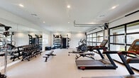 Located in Garland, Texas, this fitness center offers a comprehensively equipped exercise space designed to support a wide range of workout routines. The room features a clean, modern aesthetic with white walls, recessed lighting, and light-toned carpeted flooring that lends a bright and open atmosphere throughout.

The cardio section is well-appointed with treadmills, an elliptical machine, and a rowing machine, all positioned to take advantage of the natural light streaming in through large glass garage-style doors and windows. These expansive windows not only invite daylight into the space but also provide views of the surrounding outdoor area, enhancing the overall workout environment.

For strength training, the facility includes a full dumbbell rack with a wide selection of weights, a multi-functional cable and power rack system complete with accessories such as kettlebells, medicine balls, and resistance bands. Adjustable flat benches are conveniently distributed throughout the floor space to accommodate a variety of exercises.

Full-length mirrors line one wall, creating a sense of expanded space while allowing users to monitor their form during workouts. A set of double doors provides easy entry and exit, and the layout is thoughtfully designed to allow ample circulation space between equipment. The space delivers a functional and motivating environment suitable for a broad spectrum of fitness activities.