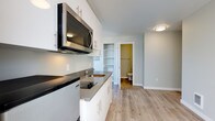 This thoughtfully designed single-floor space presents a clean, contemporary aesthetic with light hardwood-style flooring and a neutral color palette flowing throughout. The kitchen is a standout feature, outfitted with crisp white cabinetry, dark countertops, a stainless steel refrigerator, built-in cooktop, over-the-range microwave, and a single-basin sink — all arranged efficiently along a single wall for streamlined functionality. Adjacent to the kitchen, an open living area is bathed in natural light thanks to a generous corner window configuration that frames pleasant views of surrounding greenery and neighboring rooftops, creating an airy and welcoming atmosphere. A built-in shelving nook provides convenient storage just off the main living space. The bathroom is well-appointed with a subway-tiled shower and tub combination, a white vanity with sink, open wall shelving, and accessibility-friendly grab bars, all finished with consistent hardwood-style flooring. An additional room, suitable as a bedroom or flex space, benefits from a large window that draws in abundant natural light, complemented by a wall-mounted heating unit. The overall design of this space prioritizes clean lines, practicality, and a bright, modern ambiance.