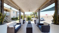 This inviting outdoor community space is thoughtfully designed for leisure and social gatherings in a warm desert climate. The centerpiece is a generously sized covered patio lounge, featuring a textured ceiling with dual ceiling fans to ensure comfortable airflow. The lounge is furnished with contemporary wicker seating accented by deep blue cushions, arranged symmetrically around low-profile coffee tables and ottomans. Tall white planters with striking desert plants add a bold, modern touch, while recessed lighting and open-air column framing allow natural light to filter through from all sides. A wall-mounted flat-screen television anchors one end of the covered lounge, making it well-suited for both casual relaxation and communal viewing. Adjacent to the lounge, a lush green lawn area provides a welcoming retreat, complete with hanging hammocks strung between sturdy posts beneath the shade of mature trees — an ideal spot for unwinding outdoors. The surrounding landscaping features native desert plants, palm trees, and decorative rock accents that complement the arid environment. A dedicated BBQ station with multiple grills and an ash disposal unit rounds out the outdoor amenities, offering residents a convenient space for outdoor cooking and dining.