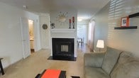 This two-floor home offers a warm and functional layout designed for comfortable everyday living. The main living area is anchored by a striking wood-burning fireplace framed by a crisp white mantel and dark surround, serving as the room's focal centerpiece. The neutral-toned carpet and soft gray walls create a calm, welcoming atmosphere throughout the space. Decorative touches on the mantel add a retro-inspired character to the room.

Adjacent to the living area, the space opens into a dining area featuring hard-surface flooring and a large window dressed with vertical blinds and sheer curtains, allowing natural light to filter through. A glimpse of an adjoining room to the left suggests additional living or utility space, with visible cabinetry hinting at a kitchen nearby.

The layout flows naturally between the main floor areas, providing a connected yet defined sense of each zone. The second floor adds additional living space, expanding the overall footprint of the home. Throughout, the design balances practicality with understated style, making it a versatile and livable residence.