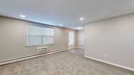 This single-floor, one-bedroom, one-bathroom unit offers a functional and thoughtfully designed layout across a compact yet efficient footprint. The combined kitchen and living room serves as the heart of the space, with the living area featuring plush wall-to-wall carpeting, neutral gray-toned walls, recessed lighting, and a window unit for climate control alongside baseboard heating. A private entrance door with recessed overhead lighting adds a welcoming entry point. The kitchen is a standout feature, showcasing dark shaker-style cabinetry, a teal glass tile backsplash, light tile flooring, and a full suite of stainless steel appliances including a gas range, over-the-range microwave, dishwasher, and refrigerator. The bedroom is a comfortable, well-sized room with soft carpeting, a window that invites natural light, a wall-mounted AC unit, baseboard heating, and a double closet for ample storage. The bathroom is finished with a dark vanity with brushed gold hardware, gray tile flooring, and a tub-shower combination accented by floor-to-ceiling striped tile surround. A hallway connects the rooms, completing this clean and move-in ready layout.