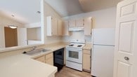 This single-floor space includes a well-appointed kitchen that serves as a central feature. The kitchen is designed with light wood cabinetry providing ample storage both above and below the counter. The layout follows an efficient L-shaped configuration, maximizing workspace and ease of movement. A stainless steel sink is positioned along the counter, complemented by light-toned laminate countertops that run continuously throughout. The kitchen is equipped with a full suite of appliances, including a white electric range with coil burners, a black dishwasher, and a full-sized white refrigerator. An overhead range hood is installed above the stove for ventilation. One of the more notable architectural elements is the vaulted ceiling above the kitchen, which adds a sense of vertical space and openness to the area. The flooring is finished in a light wood-toned laminate, contributing to the bright, neutral palette maintained throughout. The overall ambiance is clean, functional, and well-lit, offering a practical and comfortable living environment suited to everyday use.