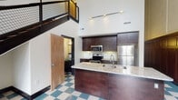Located at 245 S Liberty St in the heart of downtown Winston-Salem, NC, this distinctive two-floor space blends historic architectural character with modern updates. The main level greets visitors with striking checkerboard tile flooring in teal, cream, and charcoal tones that runs throughout, complemented by bold black trim and soaring ceilings that amplify the sense of volume and openness. Ornate decorative wall panels and tall windows with views of the surrounding cityscape lend a historic, grand ambiance to the main living area.

A sleek, modern kitchen features rich dark-espresso cabinetry, granite countertops, a central island with an undermount sink, and a full suite of stainless steel appliances including a range, over-the-range microwave, and refrigerator. Track lighting illuminates the kitchen, which also benefits from the dramatic ceiling height above.

A striking black metal staircase with grid-panel railings and warm wood treads connects the main level to the upper floor, where a mezzanine-style overlook adds architectural depth. Bathrooms are clean and functional, each featuring a tub-shower combo with brushed nickel fixtures and grab bars. Additional rooms offer flexible space with ceiling fans and transom windows that allow natural light to filter through.