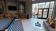 Located on Minglewood Lane in Dallas, TX, this single-story community clubroom presents a contemporary design characterized by soaring double-height ceilings and expansive floor-to-ceiling windows that flood the interior with natural light. Large-format light tile flooring spans the open layout, complemented by a bold black-and-white geometric area rug that anchors the central lounge area.

The main lounge features a plush gray sectional sofa with warm accent pillows and a sleek white coffee table, positioned adjacent to a dramatic built-in entertainment niche clad in dark stone tile with recessed lighting and a wall-mounted flat-screen TV. A cozy seating alcove with a leather sofa is integrated into this niche, creating an intimate retreat within the larger space.

High-top bistro tables with modern rattan and wire-frame bar stools are positioned near the floor-to-ceiling glass doors, offering direct views of and access to the outdoor pool courtyard. A pool table adds a recreational focal point, while decorative wall art, including a sculptural piece, enhances the modern aesthetic throughout.

A separate work or meeting area includes a wooden communal table with modern black chairs and overhead recessed lighting. The architecture features bold structural columns, curved wall elements, and a neutral gray palette that ties the entire space together into a cohesive, upscale communal environment.