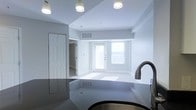 This single-floor space showcases a clean, modern design with an open-concept layout that seamlessly connects the kitchen and living areas. The kitchen is equipped with a dark granite or stone countertop and a brushed bronze gooseneck faucet over a stainless steel sink, providing a stylish contrast against the light surroundings. Recessed lighting and flush-mount ceiling fixtures illuminate the space efficiently overhead.

The living area benefits from abundant natural light streaming through two large windows flanking a white exterior door, creating a bright and airy atmosphere. The floors in the living area appear to be light-colored carpet, while the kitchen features a darker, smooth flooring surface, visually distinguishing the two zones. Neutral gray and white walls throughout contribute to the space's clean, contemporary aesthetic.

A white paneled door leads to an additional room or closet, offering functional storage or private space. The ceiling features a subtle stepped or tray design, adding a degree of architectural interest. A visible HVAC vent ensures climate control. Overall, the space presents a well-lit, minimalist environment well-suited for everyday living.