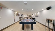 Located at 803 Village Dr in Port Coquitlam, BC, this generously sized amenity space offers a well-rounded environment designed for recreation and social gatherings. The single-floor layout is finished with light wood-look flooring throughout, crisp white walls, and recessed pot lighting that creates a bright and modern atmosphere.

The games room serves as a central highlight, featuring a full-size pool table, shuffleboard table, foosball table, and additional arcade-style games. Framed artwork and wall-mounted speakers contribute to the energetic ambiance of this area.

Adjacent to the games room is a stylish lounge area with leather sofas, area rugs, and floor-to-ceiling windows that invite an abundance of natural light and provide views of the surrounding outdoor courtyard and greenery.

The kitchen and dining area is equally impressive, showcasing a large marble-topped island with bar seating, warm wood cabinetry with open shelving, pendant lighting, and a wall-mounted TV. A full-size refrigerator and additional counter space support hosting and catering needs.

A separate dining zone accommodates multiple large tables with ample seating, complemented by upholstered banquette seating and access to an outdoor area with barbecue facilities. A washroom completes the space.
