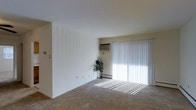 This single-floor residential space offers a clean, functional layout designed for comfortable everyday living. The main living area is a standout feature, characterized by its open, airy feel and neutral color palette throughout. Smooth white walls and a light-toned ceiling create a bright, welcoming atmosphere that is further enhanced by an expansive sliding glass door fitted with vertical blinds. This large door allows abundant natural light to flood the room, casting a warm glow across the wall-to-wall carpeted flooring. The carpet adds a soft, comfortable underfoot feel to the space and contributes to the overall understated aesthetic. Climate control is provided by a wall-mounted air conditioning unit positioned near the sliding door, ensuring year-round comfort. A baseboard heating unit is also visible along the exterior wall, offering an additional layer of temperature regulation. The living space is generously proportioned, providing ample room for a variety of furniture arrangements. A tall indoor plant near the glass door adds a touch of natural greenery to the otherwise minimalist interior. The overall ambiance is simple, bright, and practical, making this a versatile space well-suited for a range of living arrangements.