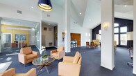 This single-floor space presents a well-appointed communal lounge or clubroom with a striking architectural design. The most notable feature is the dramatically vaulted ceiling, which amplifies the sense of openness and volume throughout the main area. Crisp white walls contrast beautifully with deep navy blue carpeting, establishing a refined color palette carried throughout the space.

The main lounge area is anchored by a cluster of warm tan leather armchairs arranged around a sleek glass-top coffee table, accented with a white orchid centerpiece. A bold navy pendant light fixture overhead adds a contemporary focal point. Structural white columns define the open floor plan while subtly separating distinct functional zones.

Toward the rear, a bar-height counter with blue stools serves as a casual dining or social area, while a separate seating nook furnished with yellow-toned chairs and a dining table offers additional gathering space. Recessed lighting supplements natural light filtering through multiple windows. Gold wall-mounted sconces and decorative brass accents add warmth and elegance, completing the polished, hospitality-inspired ambiance of the space.
