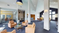 This single-floor space presents a well-appointed communal lounge or clubroom with a striking architectural design. The most notable feature is the dramatically vaulted ceiling, which amplifies the sense of openness and volume throughout the main area. Crisp white walls contrast beautifully with deep navy blue carpeting, establishing a refined color palette carried throughout the space.

The main lounge area is anchored by a cluster of warm tan leather armchairs arranged around a sleek glass-top coffee table, accented with a white orchid centerpiece. A bold navy pendant light fixture overhead adds a contemporary focal point. Structural white columns define the open floor plan while subtly separating distinct functional zones.

Toward the rear, a bar-height counter with blue stools serves as a casual dining or social area, while a separate seating nook furnished with yellow-toned chairs and a dining table offers additional gathering space. Recessed lighting supplements natural light filtering through multiple windows. Gold wall-mounted sconces and decorative brass accents add warmth and elegance, completing the polished, hospitality-inspired ambiance of the space.