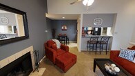 This single-floor residence features an open-concept layout that integrates the living, dining, and kitchen areas into one cohesive space. The living area is anchored by a fireplace on one end, complemented by neutral-toned carpet flooring that extends throughout. Bold gray walls provide a strong visual backdrop, and a ceiling fan with a light fixture overhead serves both functional and aesthetic purposes.

A plush red armchair with a matching ottoman serves as the focal point of the living area, positioned alongside a dark coffee table. The space flows naturally into a dining nook in the background, where a compact bistro-style table and bar stools create an intimate eating area. Wall décor adds a personalized touch to this section of the room.

To the right, a raised kitchen bar with barstool seating connects the kitchen to the main living area. The kitchen features dark cabinetry and appliances visible beyond the bar counter, with a wall clock mounted nearby. The open-concept design ensures seamless communication between all areas, making the space feel connected and functional while maintaining a consistent color palette and warm ambiance throughout.