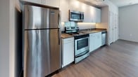 Located at 1024 NE 7th St in Oklahoma City, Oklahoma, The Seven Model 1 is a single-floor residence designed with a clean, contemporary aesthetic. The layout flows logically between living, kitchen, and bathroom areas, creating a functional and comfortable environment.

The kitchen is a standout feature, outfitted with stainless steel appliances including a full-size refrigerator, electric range, over-the-range microwave, and dishwasher. White cabinetry extends to the upper walls, providing generous storage, while dark countertops offer a sharp contrast. Wood-look plank flooring runs throughout the main living areas, lending warmth and a modern feel to the space.

The living area is open and bright, with natural light entering through windows that frame views of the surrounding neighborhood. Clean white walls and neutral tones create a versatile backdrop, while the open layout offers flexibility in furniture arrangement.

The bathroom features light tile flooring, built-in wall shelving for storage, and a walk-in style shower unit. The space is practical and well-organized, with a layout that maximizes utility. Overall, The Seven Model 1 presents a straightforward, modern living environment suited for those seeking a well-appointed single-level space in Oklahoma City.
