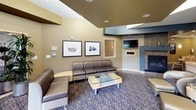 This single-floor communal space is thoughtfully designed to serve as both a relaxing retreat and a functional gathering area. The layout is divided into several distinct zones, each serving a specific purpose while maintaining a cohesive aesthetic throughout.

The main lounge area features plush brown leather seating arranged around a glass-topped coffee table, anchored by a stone fireplace and a wall-mounted flat-screen television. Decorative framed artwork adorns the textured walls, complemented by warm, recessed lighting and layered ceiling details that add architectural depth.

Adjacent to the lounge, a casual dining area offers multiple tables with light wood and green upholstered chairs, ideal for small group gatherings or individual use. Framed photographic prints with serene water and boat imagery lend a tranquil, nature-inspired character to this section.

The space opens up further into a bright, expansive area bathed in natural light through large floor-to-ceiling windows that frame lush outdoor greenery. Tall indoor plants and additional seating clusters create an inviting, airy atmosphere. Structural columns with warm wood paneling add visual interest and define separate seating pockets.

A connecting corridor with hardwood flooring and pendant lighting leads to an additional area with a reception-style counter, decorative mirror, and accent furnishings. The patterned carpeting flows consistently throughout, tying the entire space together in a polished, welcoming environment.