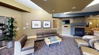 This single-floor communal space is thoughtfully designed to serve as both a relaxing retreat and a functional gathering area. The layout is divided into several distinct zones, each serving a specific purpose while maintaining a cohesive aesthetic throughout.

The main lounge area features plush brown leather seating arranged around a glass-topped coffee table, anchored by a stone fireplace and a wall-mounted flat-screen television. Decorative framed artwork adorns the textured walls, complemented by warm, recessed lighting and layered ceiling details that add architectural depth.

Adjacent to the lounge, a casual dining area offers multiple tables with light wood and green upholstered chairs, ideal for small group gatherings or individual use. Framed photographic prints with serene water and boat imagery lend a tranquil, nature-inspired character to this section.

The space opens up further into a bright, expansive area bathed in natural light through large floor-to-ceiling windows that frame lush outdoor greenery. Tall indoor plants and additional seating clusters create an inviting, airy atmosphere. Structural columns with warm wood paneling add visual interest and define separate seating pockets.

A connecting corridor with hardwood flooring and pendant lighting leads to an additional area with a reception-style counter, decorative mirror, and accent furnishings. The patterned carpeting flows consistently throughout, tying the entire space together in a polished, welcoming environment.