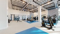 Located at 5 Port Imperial Blvd in Weehawken, NJ, this professionally appointed fitness center offers a spacious, single-level facility designed to support a comprehensive range of workout routines. The main gym floor is an open, airy space characterized by soaring ceilings fitted with industrial-style grid frameworks, pendant lighting, and ceiling fans that enhance airflow and ambiance. Large floor-to-ceiling windows line portions of the perimeter, flooding the space with abundant natural light and offering views of the surrounding area. The flooring is a combination of light-toned resilient surfaces and vibrant blue-accented zones that visually define different workout areas. The main floor is equipped with a full array of Technogym cardio machines—including ellipticals, stationary bikes, and steppers—alongside strength equipment, selectorized machines, free weights, and squat racks. A prominent blue mosaic tile accent wall adds a bold, modern design element throughout. Built-in wood cabinetry and a water station provide added convenience. A separate studio room features warm hardwood flooring, full-length mirrors, recessed cove lighting, a large display screen, ballet barres, and spin bikes, making it ideal for group fitness and mind-body classes. The overall ambiance is clean, modern, and energizing.
