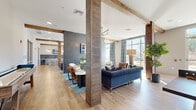 Located at 1804 Appaloosa Ln in Buford, GA, this single-floor community space offers a generously sized, open-concept layout that seamlessly blends a living area with a kitchen lounge. The interior design strikes a sophisticated balance between modern and rustic, highlighted by prominent wood-wrapped structural columns and exposed beam accents that frame each zone with warmth and character.

The living area features multiple seating arrangements, including a tufted navy sofa, leather accent chairs, and a channeled leather couch, all anchored by teal area rugs and complemented by bold artwork on the walls. A linear fireplace set against a large-format gray tile accent wall serves as a striking focal point, topped by a wall-mounted television and illuminated by a sculptural brass and globe pendant chandelier.

The kitchen area includes a clean white countertop island with bar seating, a geometric dark tile backsplash, under-counter beverage refrigeration, pendant lighting, and an additional wall-mounted television. A game area with an air hockey table adds recreational appeal.

Throughout the space, wide-plank wood-look flooring, floor-to-ceiling windows dressed with plaid drapery, and abundant natural light create a bright, welcoming ambiance with open views of the surrounding landscape.
