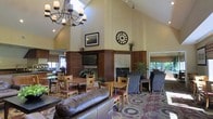 This single-floor communal lounge offers a warm and inviting atmosphere defined by its dramatically vaulted ceilings, which lend a spacious, airy quality to the interior. A striking wrought-iron chandelier hangs prominently overhead, complementing the rustic yet refined aesthetic of the space. At the heart of the room stands a wood-paneled fireplace surround with a tiled hearth, above which a mounted flat-screen television is positioned for easy viewing. A large decorative clock and framed artwork adorn the walls, adding character to the space.

The seating is thoughtfully arranged to accommodate a variety of uses. Casual dining-style tables with wooden chairs featuring cushioned seats are grouped near the windows, while upholstered lounge chairs and a leather sofa provide comfortable areas for relaxing. Patterned carpeting in rich tones anchors the seating areas and contributes to the room's cozy ambiance. Lush potted plants are placed throughout, adding greenery and life to the interior. Multiple windows along the perimeter allow abundant natural light to filter in, enhancing the welcoming character of this well-appointed shared living space.